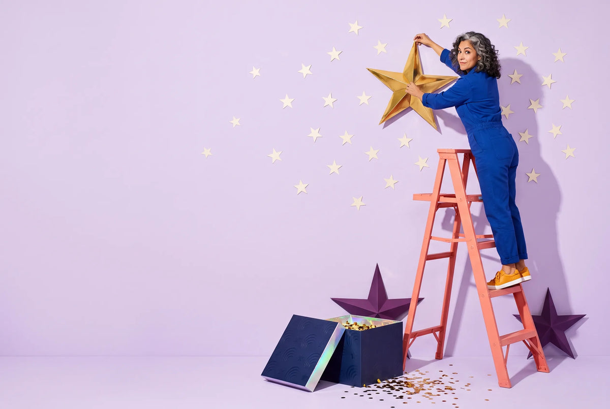 A woman in a blue jumpsuit on a coral stepladder pins a large gold paper star to a lavender wall, with an open navy Everwow gift box below