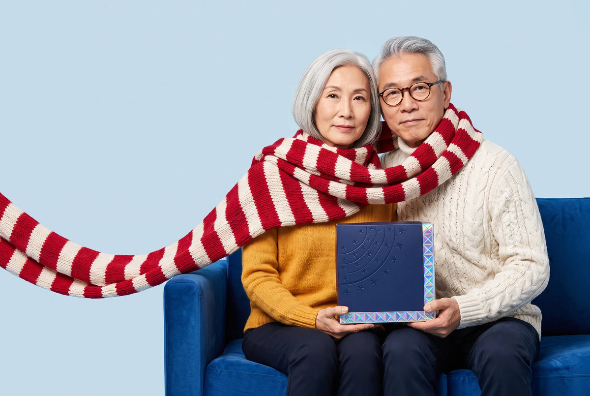 An older couple wrapped together in one long red and white striped scarf, holding a navy Everwow gift box on a blue velvet sofa