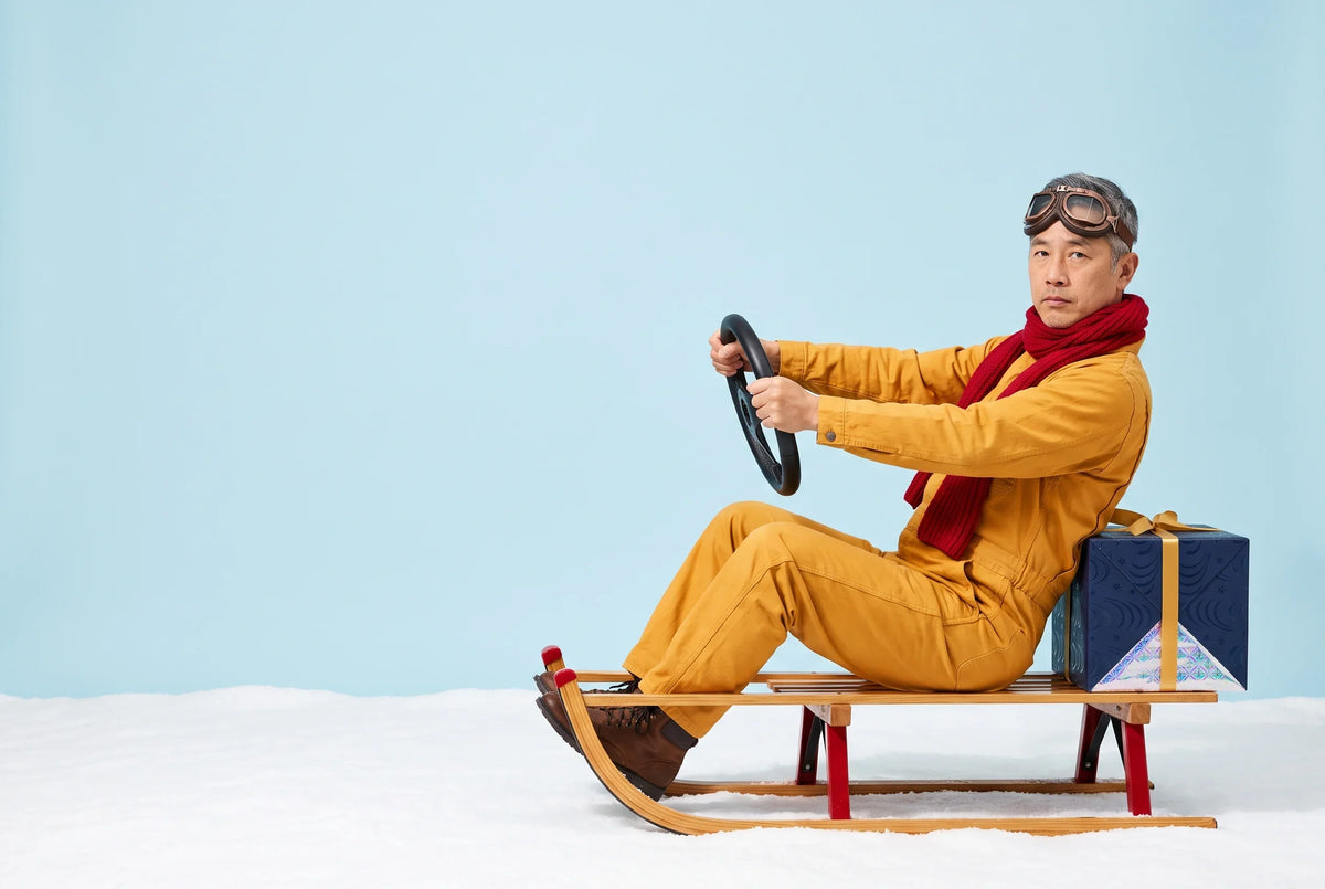 A man in a yellow jumpsuit, red scarf and aviator goggles sits in a wooden sled on snow, gripping a steering wheel, with a navy Everwow gift box tied to the back with a gold ribbon