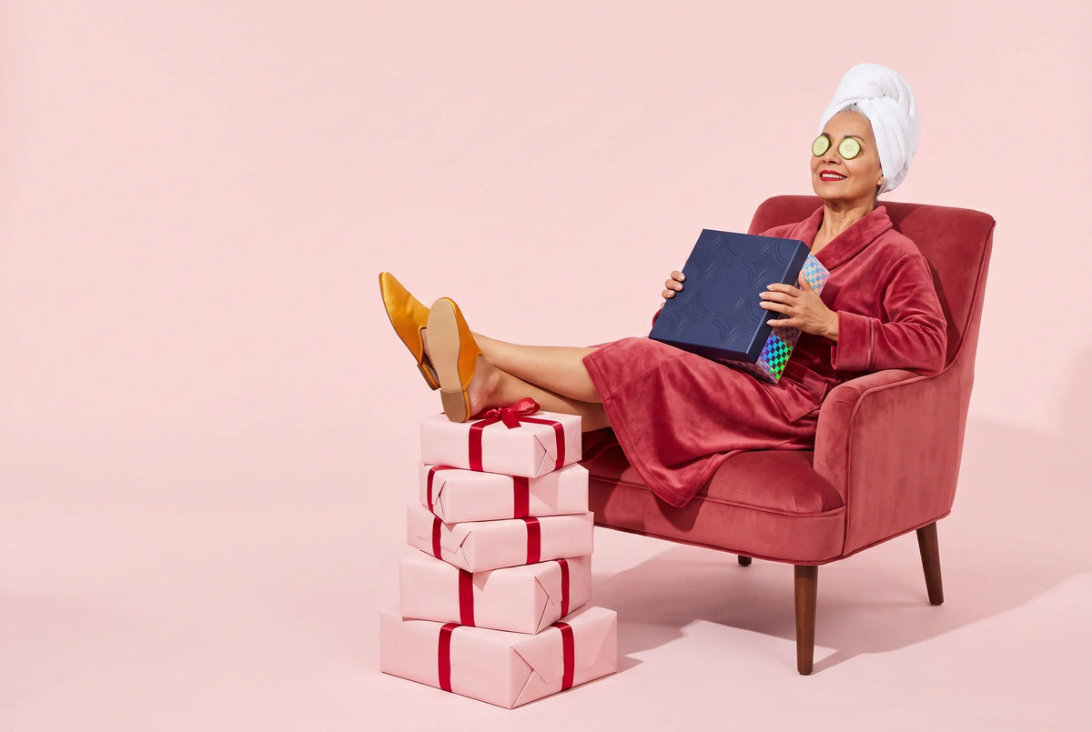 A woman in a rose-red velvet robe and white towel turban relaxes in a velvet armchair with cucumber slices over her eyes, her yellow slippers resting on a stack of pink-wrapped presents as she holds a navy Everwow gift box