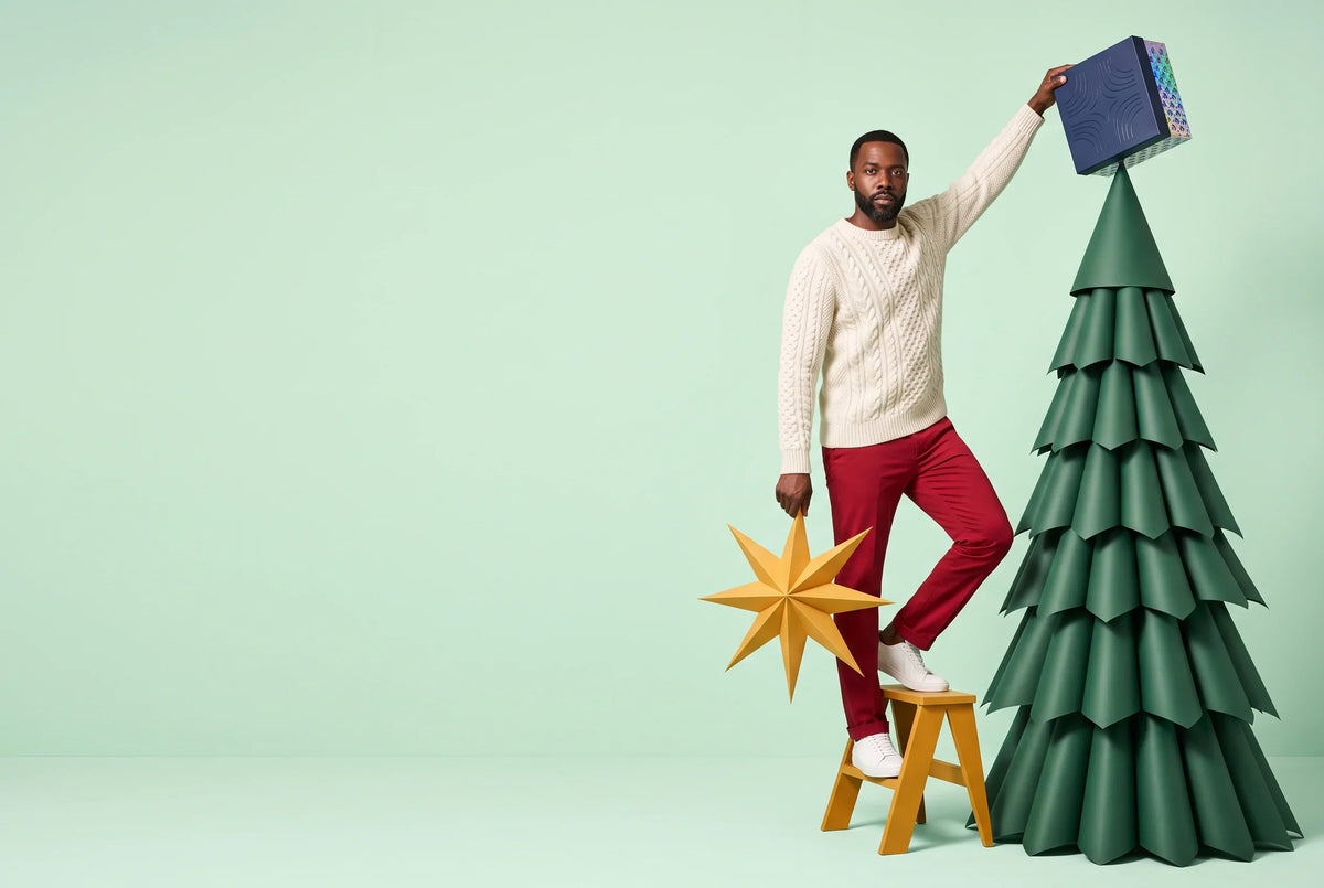 A man in an off-white cable-knit sweater and red trousers stands on a yellow step stool, placing a navy Everwow gift box on top of a green paper Christmas tree while holding a golden paper star