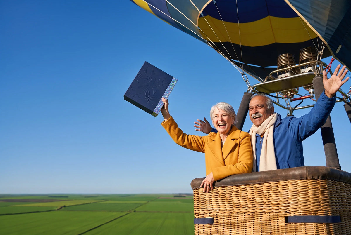 A laughing older couple in a hot air balloon basket high above green fields, the woman in a yellow coat holding up a navy Everwow gift box against a clear blue sky