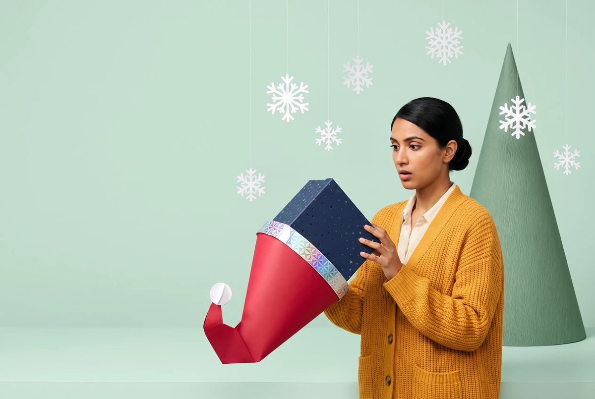 A woman in a mustard cardigan pulls a navy Everwow gift box out of a red paper Santa hat, with paper snowflakes on a pale mint set