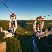 Two friends racing side by side on parallel ziplines over a forest gorge, Ziplining experience gift