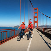 Two cyclists riding the bridge bike lane past a rust-orange tower, San Francisco Adventure experience gift