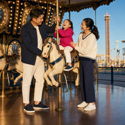 Child laughing on a carousel horse with parents alongside at evening, Theme Park Day experience gift
