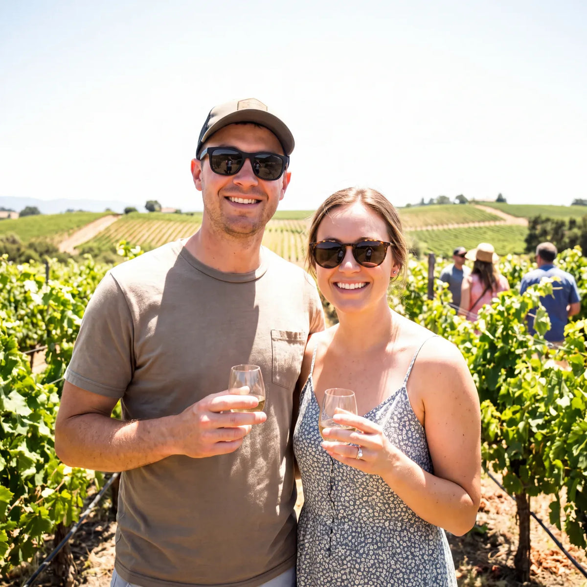 Couple in sunglasses holding glasses of white wine among the vines at a vineyard tasting