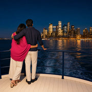 Couple at a cruise boat rail watching city lights across the harbor, City Lights Cruise experience gift