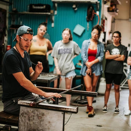 Glassblower shaping molten glass at a bench while a small class watches in the studio
