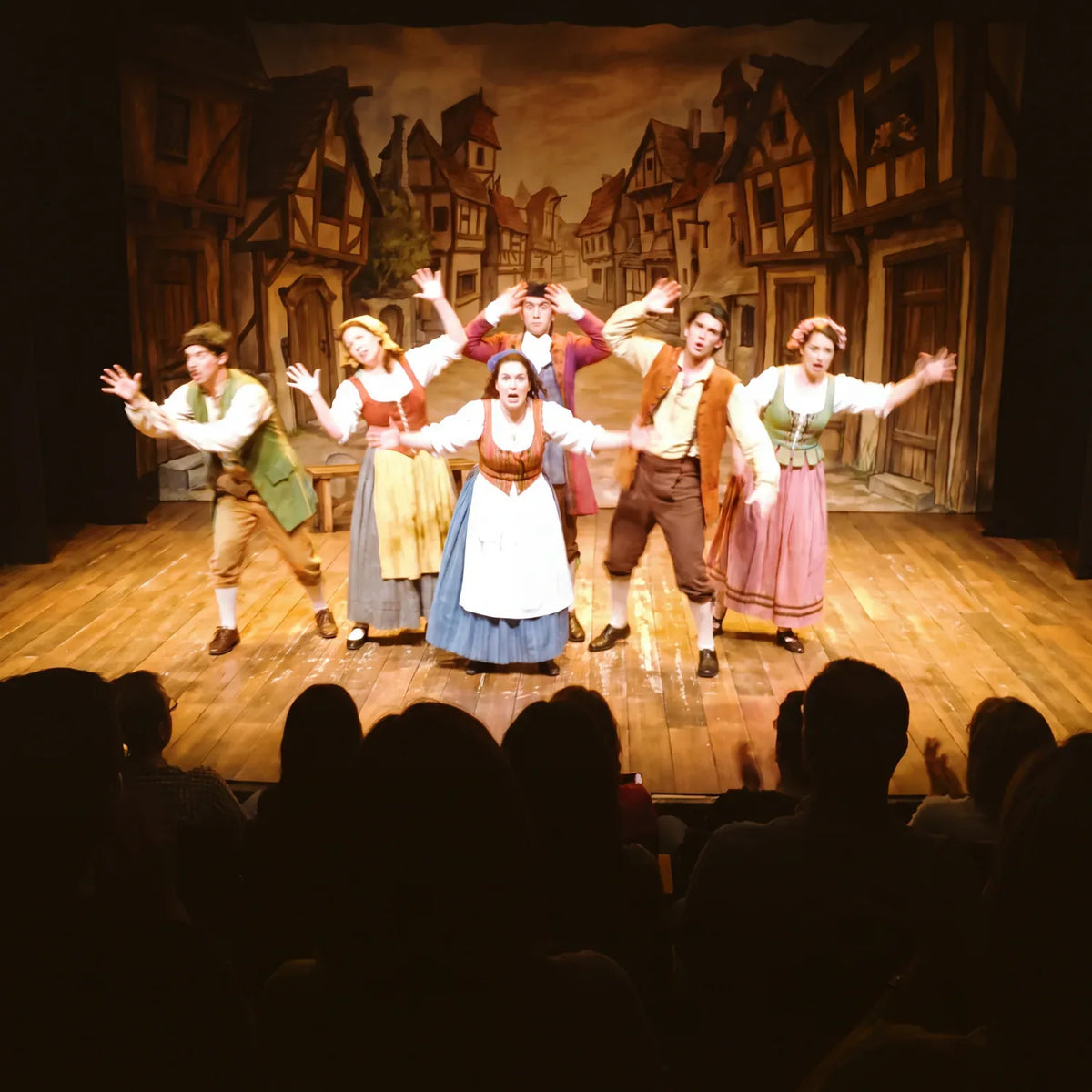 Costumed cast performing on a village-set stage in front of a seated theatre audience