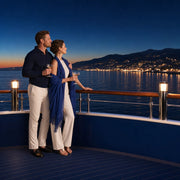 Couple with coupes at a yacht rail at dusk, coastline lights beyond, Iconic Cruises & Sailing Luxe experience gift