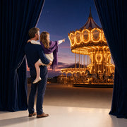 Parent lifting a child on one hip to watch a glowing carousel at dusk, Iconic Funday Luxe experience gift