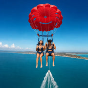 Couple flying beneath a red parasail above turquoise water, Parasailing experience gift