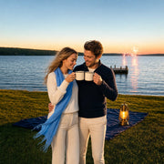 Couple toasting enamel mugs at a lakeshore picnic as distant fireworks glow, Iconic Fourth of July experience gift