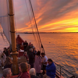 Passengers at the rail of a wooden tall ship watching a vivid orange sunset at sea
