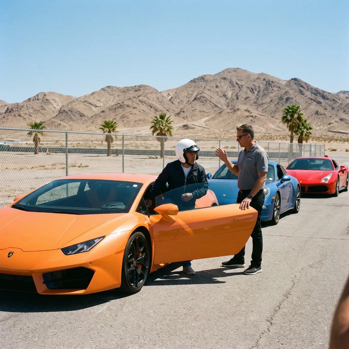 Instructor briefing a helmeted driver beside an orange supercar at a desert racetrack