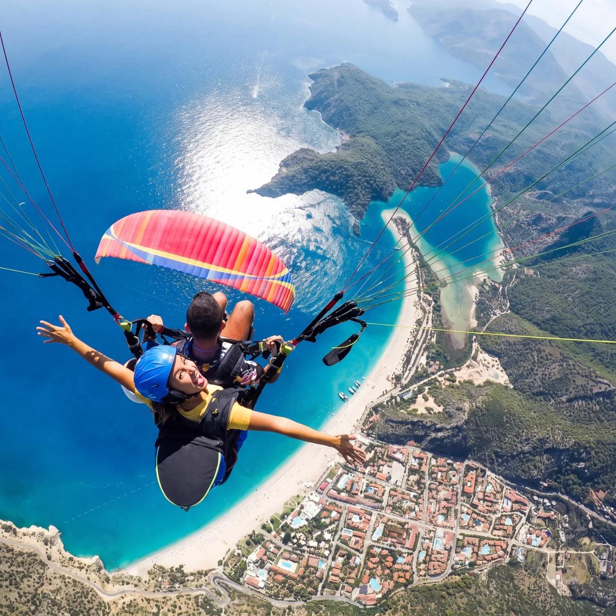 Tandem paragliding pair with arms outstretched high above a turquoise bay and coastal town