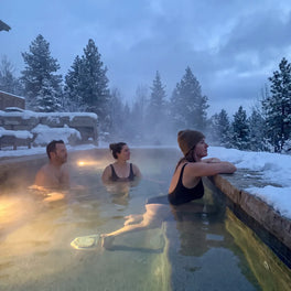 Three people relaxing in a steaming outdoor hot pool surrounded by snow-covered pines