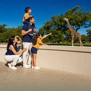 Family watching a giraffe reach into the treetops at a zoo railing, Zoo Visit experience gift